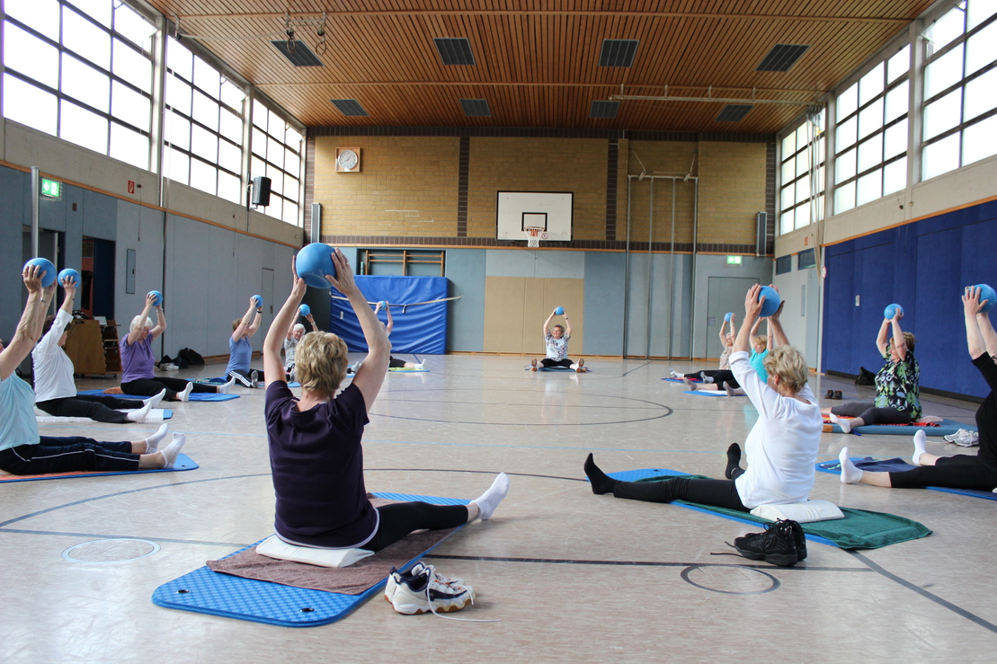 Gruppentraining in einer Turnhalle, Teilnehmer heben Medizinb&auml;lle &uuml;ber Kopf auf Matten im Halbkreis.