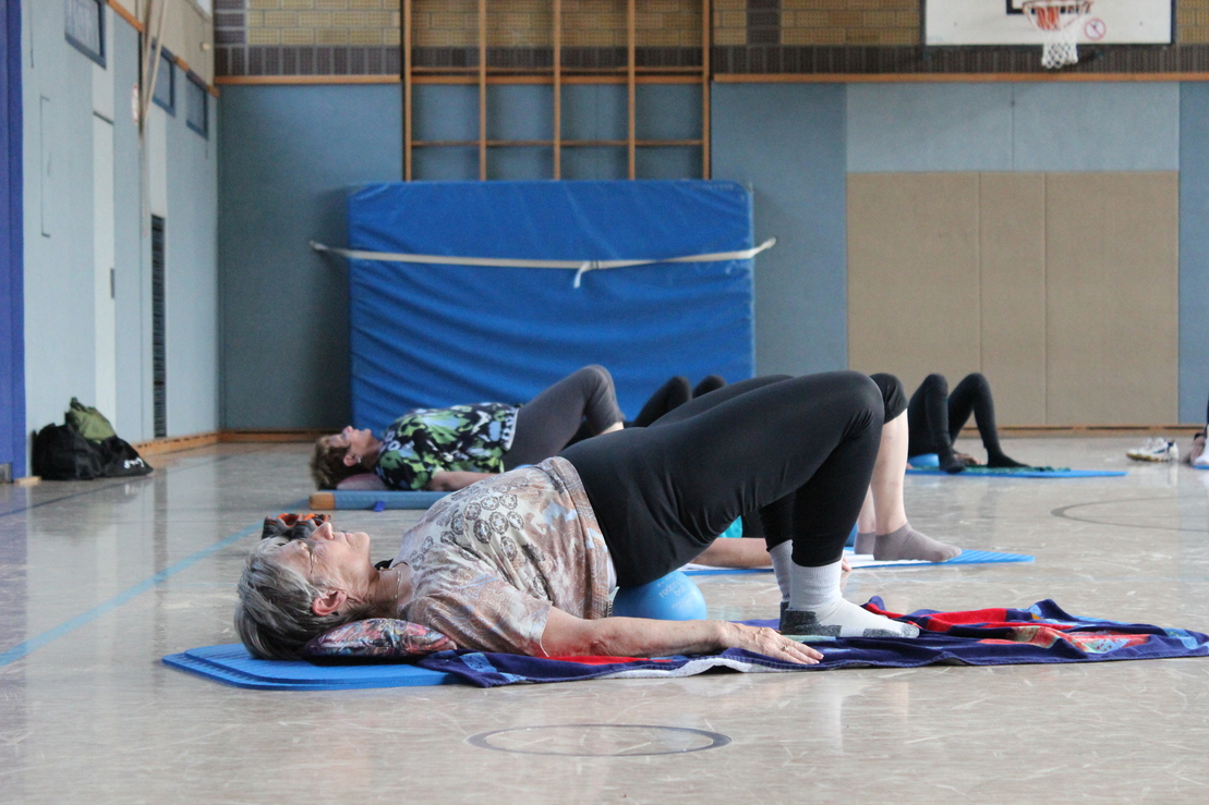 &Auml;ltere Frau macht eine Yoga-&Uuml;bung mit einem Ball auf einer Matte in einer Sporthalle, weitere Teilnehmer im Hintergrund.