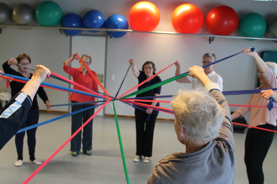Gruppe von &auml;lteren Menschen, die elastische B&auml;nder in einem Raum mit Fitnessger&auml;ten halten und gemeinsam eine &Uuml;bung machen.