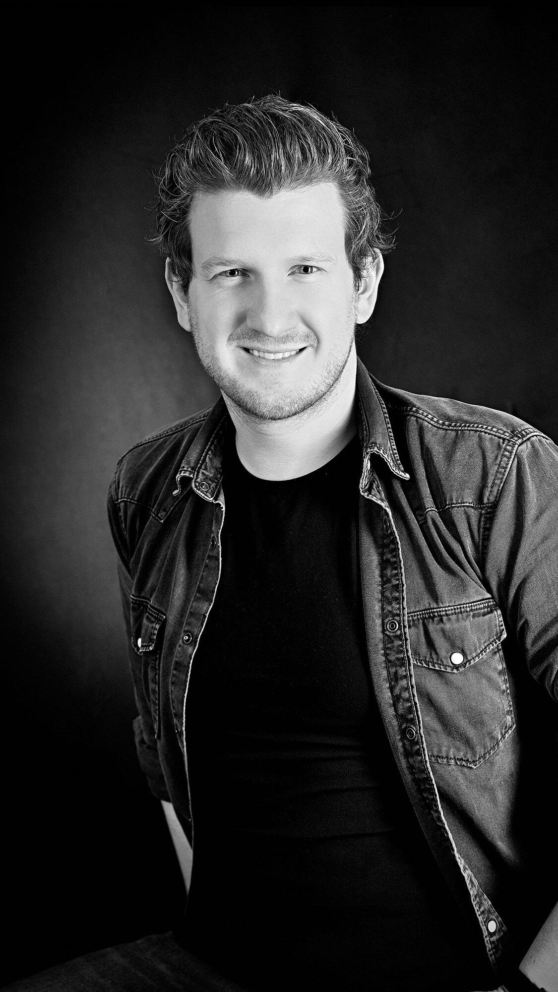 Portr&auml;t eines l&auml;chelnden Mannes in einem schwarzen Hemd und Jeansjacke vor einem dunklen Hintergrund.