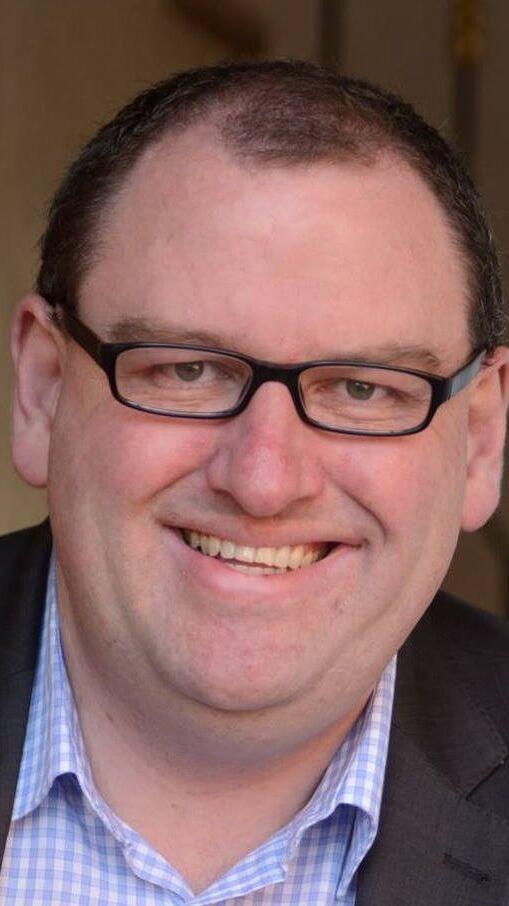 Portrait eines l&auml;chelnden Mannes mit Brille, der einen Anzug und ein kariertes Hemd tr&auml;gt. Hintergrund unscharf.