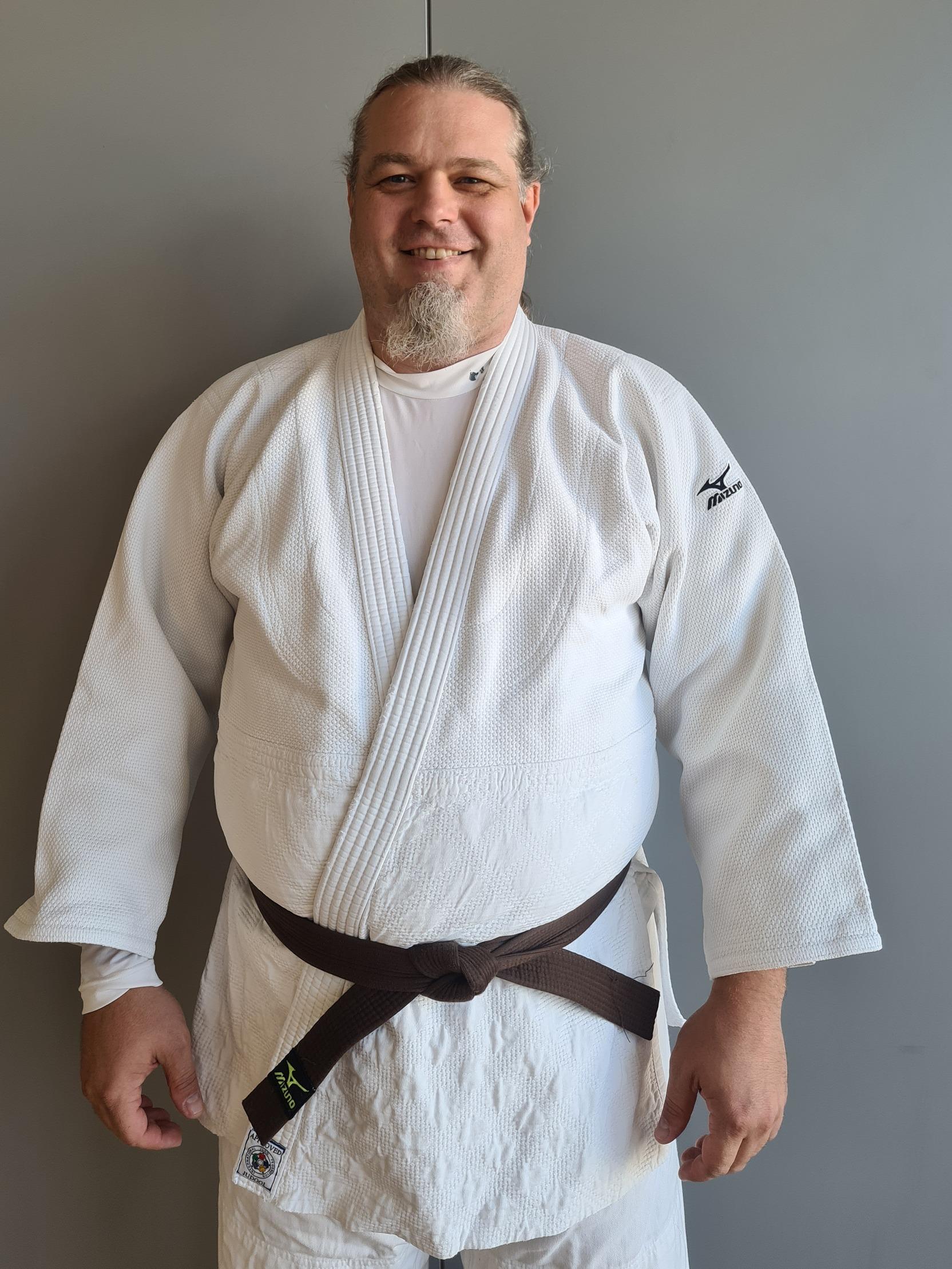 Mann in judof&auml;higer Kleidung mit braunem G&uuml;rtel steht vor einer grauen Wand und l&auml;chelt.