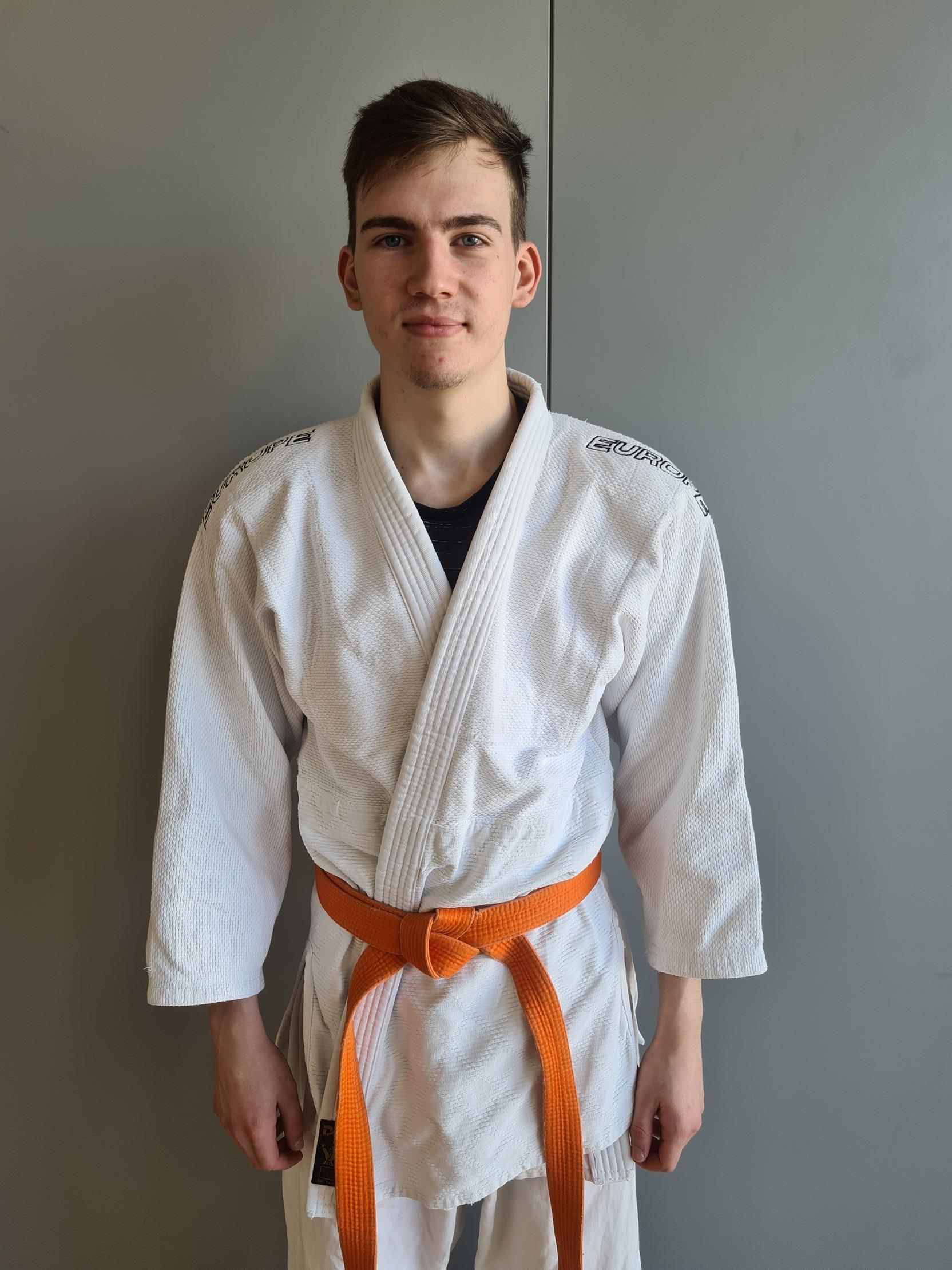 Junger Mann tr&auml;gt einen wei&szlig;en Judogi mit orangefarbenem G&uuml;rtel und steht vor einer grauen Wand.