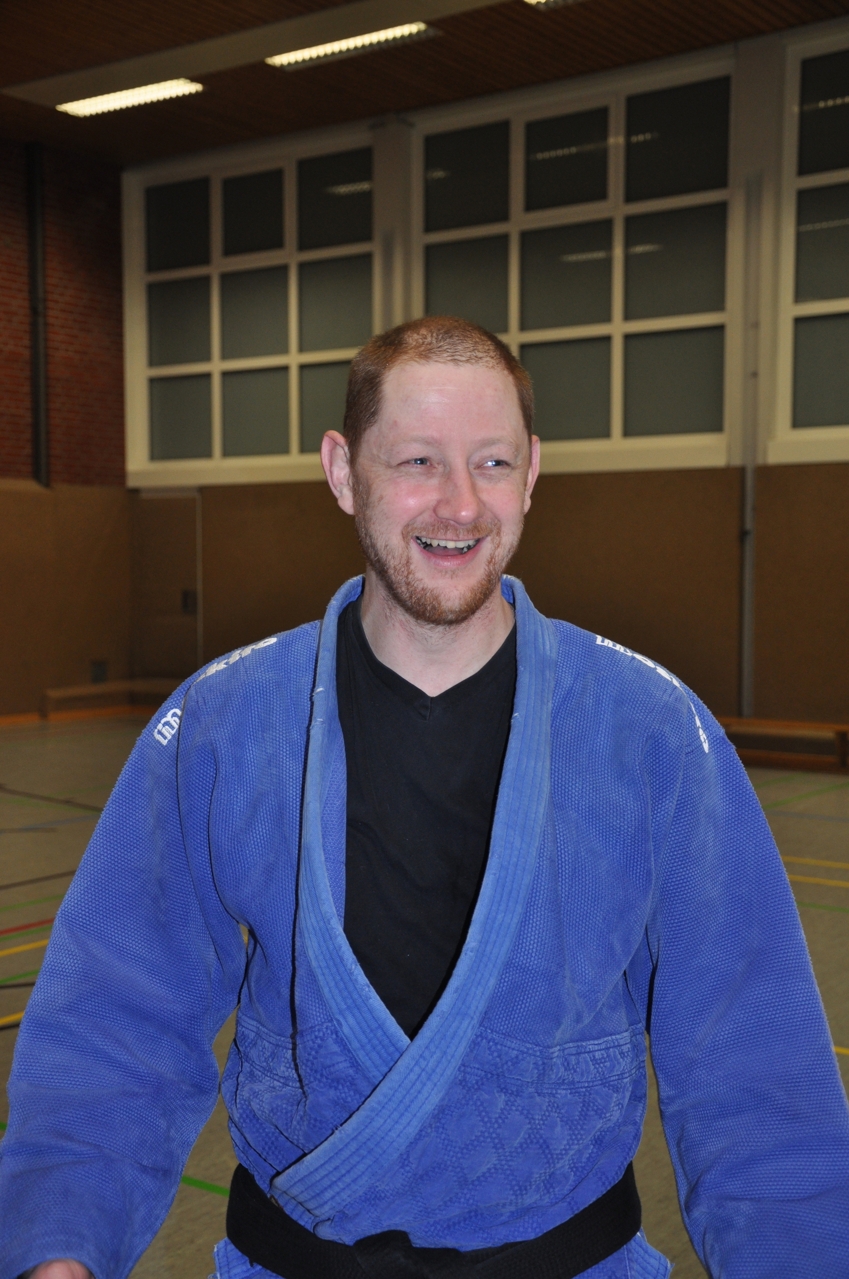 L&auml;chelnder Mann in blauer Judo-Gi, steht in einer Sporthalle mit Fensterfront im Hintergrund.