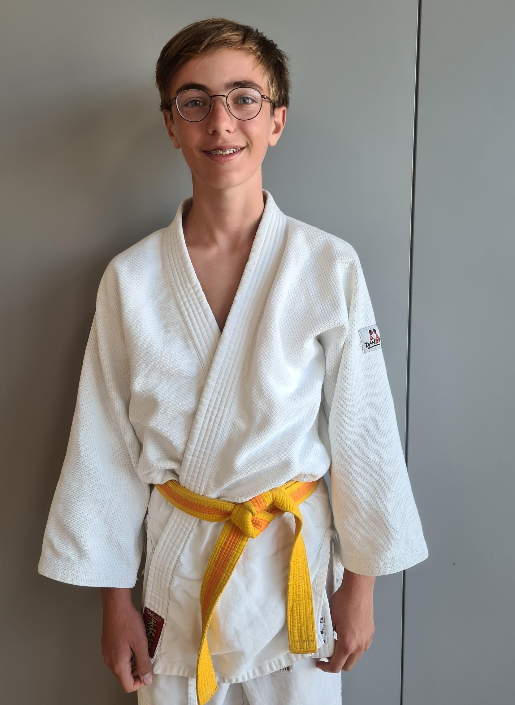 Ein junger Mann in einem wei&szlig;en Judoanzug mit gelbem G&uuml;rtel steht vor einer grauen Wand und l&auml;chelt.