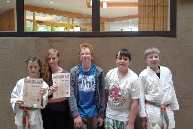 F&uuml;nf Kinder, darunter zwei in Karateanz&uuml;gen, halten Urkunden in einem hellen Raum mit gro&szlig;en Fenstern.