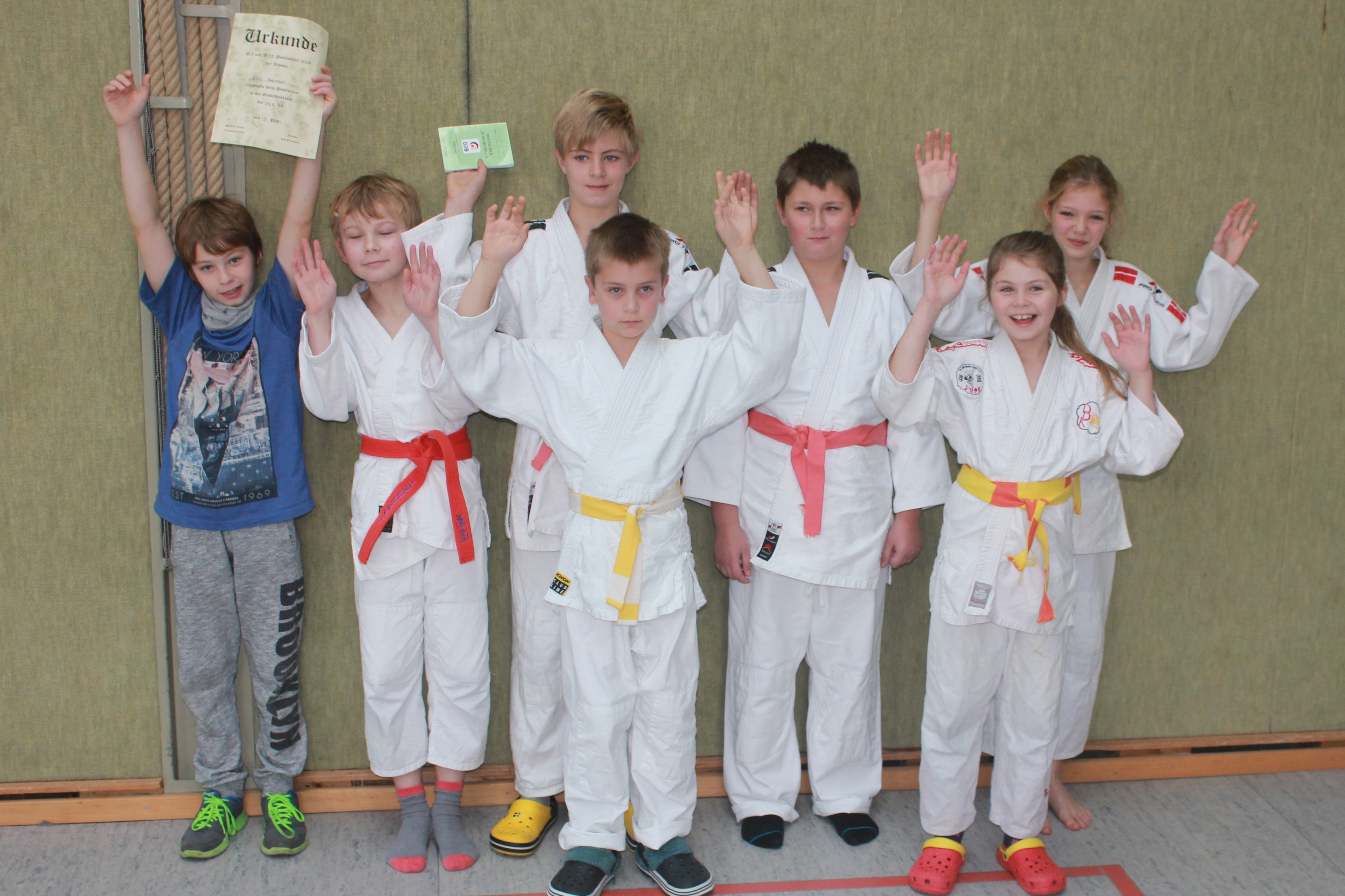 Gruppe von acht Kindern in Judoanz&uuml;gen, einige halten Urkunden und Karten, alle mit erhobenen H&auml;nden.