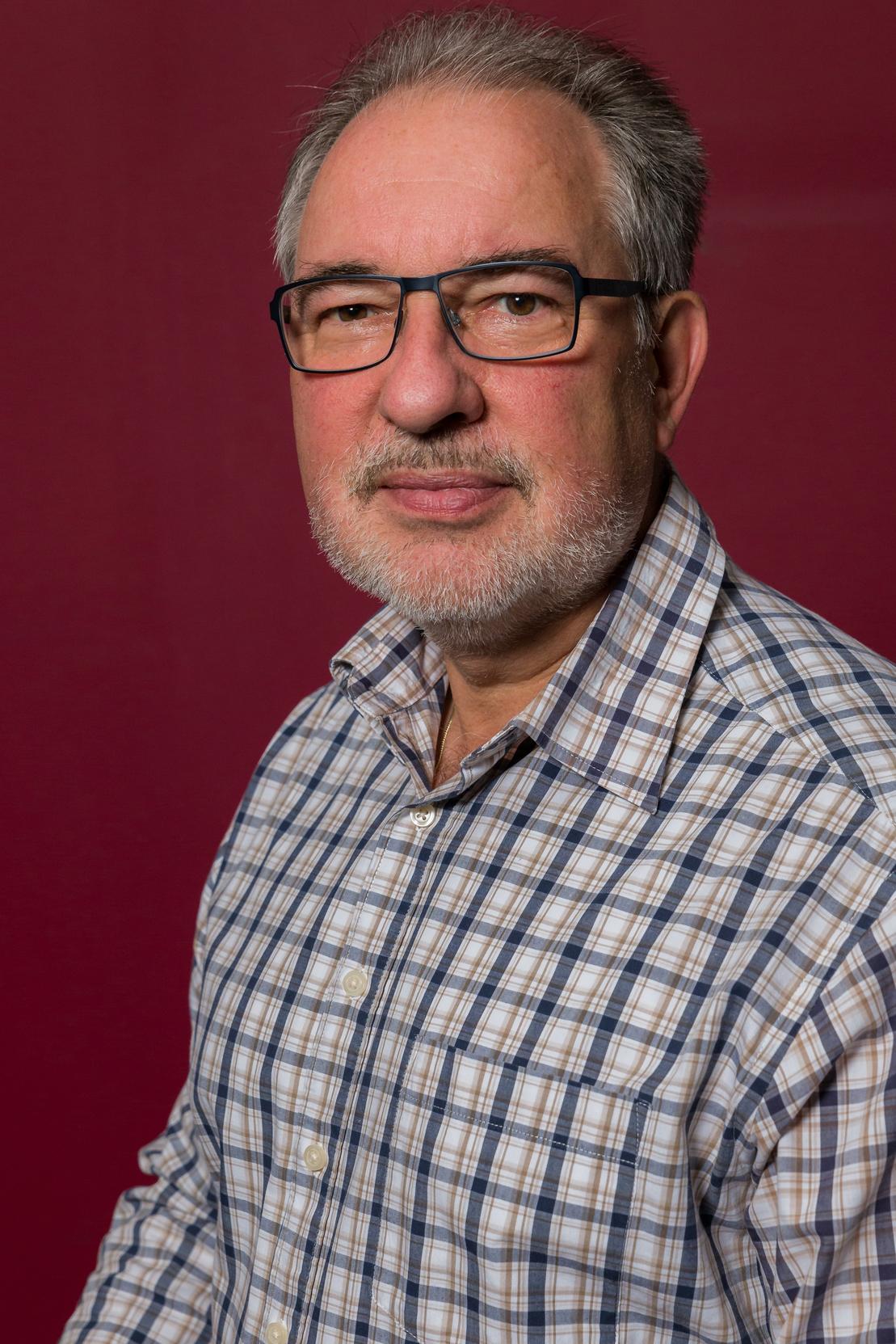 Portr&auml;t eines Mannes mit Brille und gestreifter Hemd vor einem roten Hintergrund. Er schaut direkt in die Kamera.
