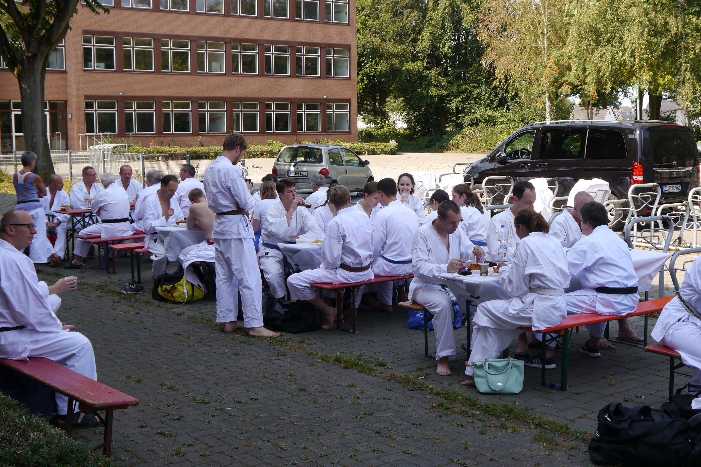 Gruppenszene von Menschen in wei&szlig;en Trainingsanz&uuml;gen, die an Tischen im Freien sitzen und essen.