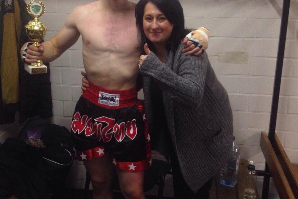 Athlet in Trainingskleidung hält einen Pokal, während eine Frau Daumen hoch zeigt, im Hintergrund ein Wasserbehälter.