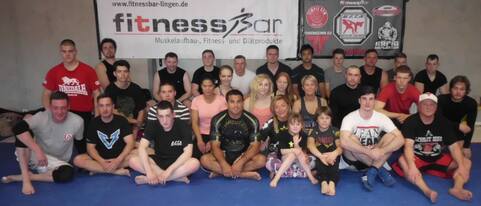 Gruppe von 30 Personen auf einer blauen Matte in einem Fitnessstudio, umgeben von Fitness- und Kampfsportwerbung.