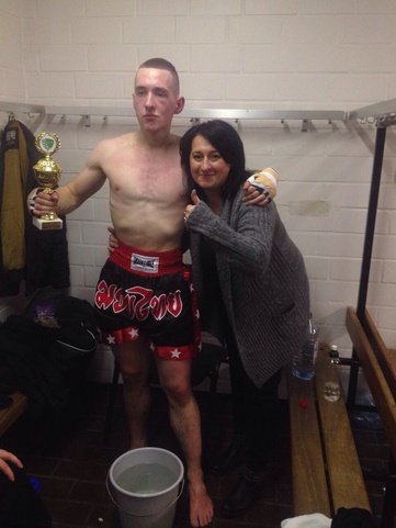 Athlet in Trainingskleidung h&auml;lt einen Pokal, w&auml;hrend eine Frau Daumen hoch zeigt, im Hintergrund ein Wasserbeh&auml;lter.