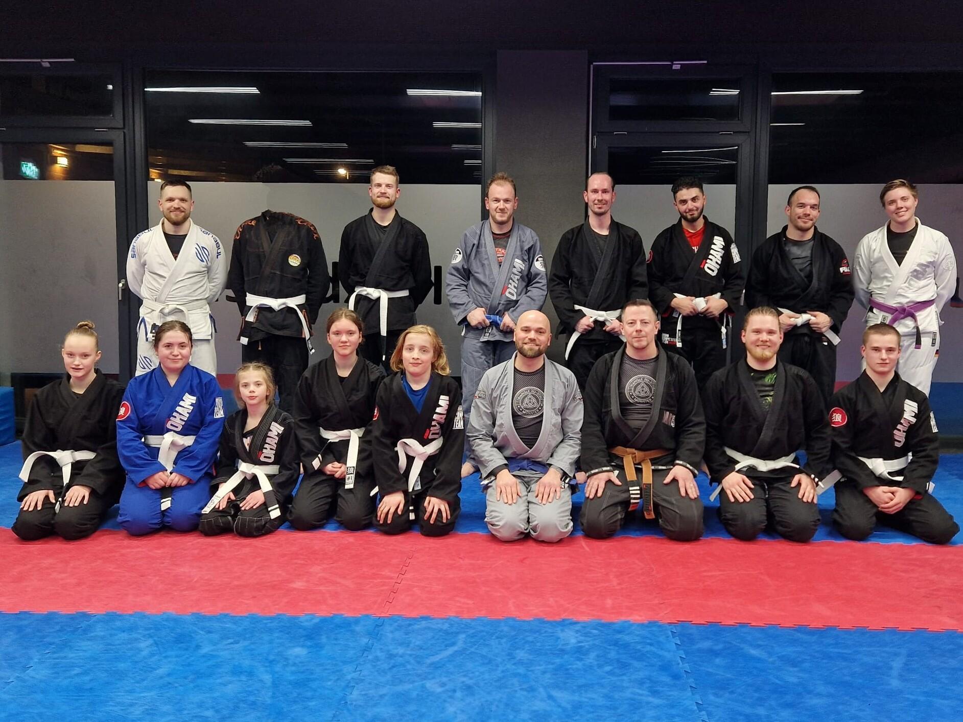 Gruppe von 15 Personen in Kampfsportanzügen auf einer Trainingsmatte, lächelnd und in verschiedenen Positionen versammelt.