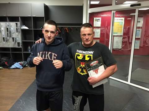 Zwei M&auml;nner stehen in einem Fitnessstudio, einer tr&auml;gt einen Nike-Hoodie und der andere ein T-Shirt mit der Aufschrift "THROWDOWN".