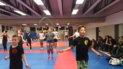 Kinder und Jugendliche springen mit Seilen in einer Sporthalle, umgeben von Zuschauern auf B&auml;nken.