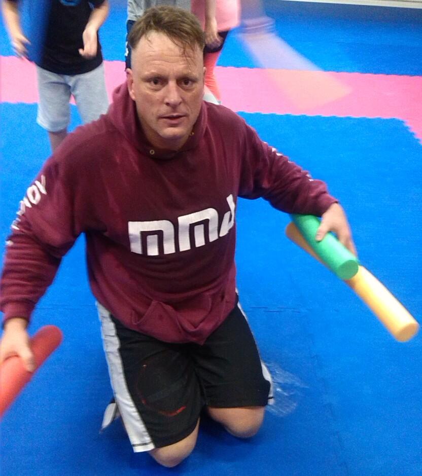 Ein Mann kniet auf einer blauen Matte und h&auml;lt zwei farbige Schaumstoffst&auml;be in den H&auml;nden, w&auml;hrend Kinder im Hintergrund spielen.