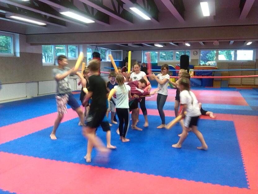 Gruppe von Kindern und Jugendlichen trainiert in einem Sportraum mit blauen und roten Matten, einige halten Schlagst&ouml;cke.