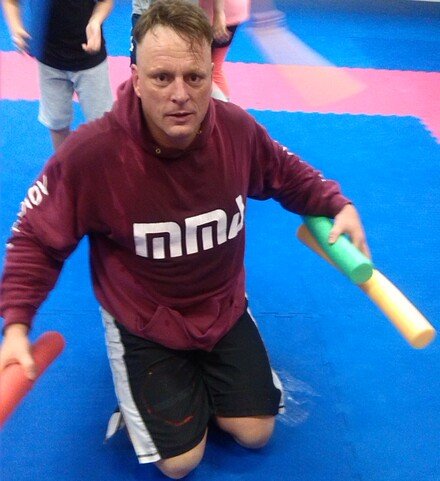 Ein Mann kniet auf einer blauen Matte und h&auml;lt zwei farbige Schaumstoffst&auml;be in den H&auml;nden, w&auml;hrend Kinder im Hintergrund spielen.