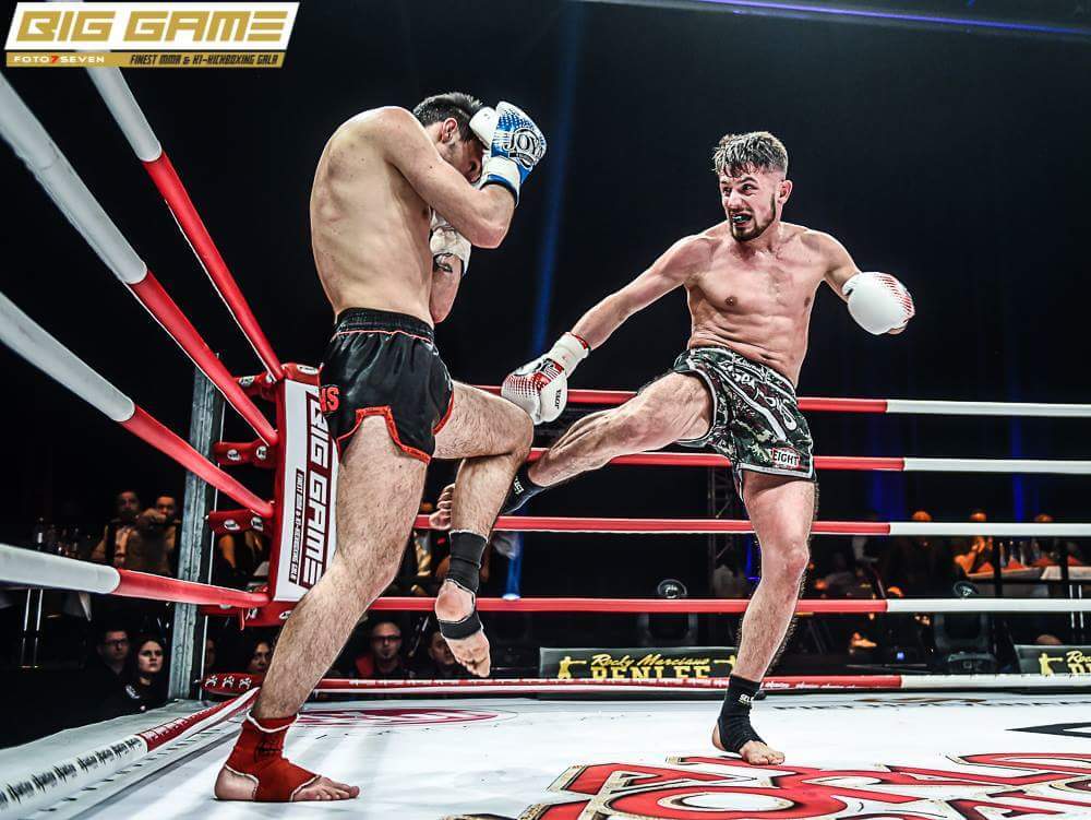 Zwei K&auml;mpfer im Boxring, einer kickt hoch, w&auml;hrend der andere sich abwehrt. Rote und wei&szlig;e Seile um den Ring.