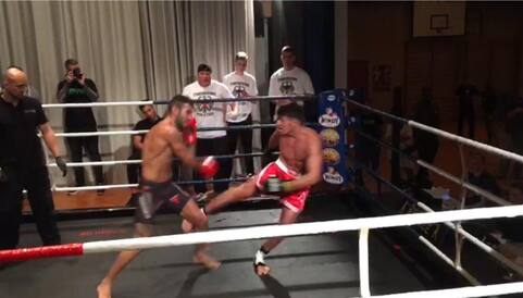 Zwei Boxer im Ring w&auml;hrend eines Kampfes, einer setzt einen Tritt an, Zuschauer beobachten gespannt im Hintergrund.