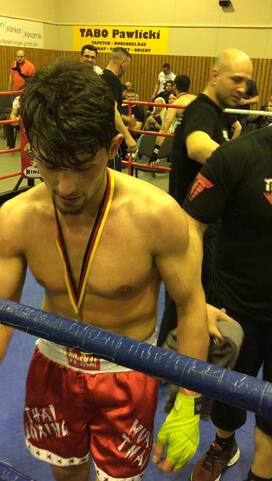 Ein junger, muskul&ouml;ser Kampfsportler in roten Shorts mit Medaille steht im Ring, w&auml;hrend Betreuer ihn umgeben.