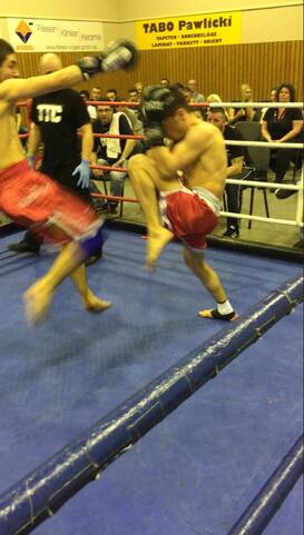 Zwei K&auml;mpfer im Ring w&auml;hrend eines Wettkampfs, einer f&uuml;hrt einen Kick aus, Zuschauer im Hintergrund.