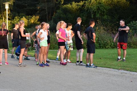 Gruppe von Jugendlichen in Sportkleidung h&ouml;rt einem Trainer im Freien aufmerksam zu. Gr&uuml;nfl&auml;che im Hintergrund.
