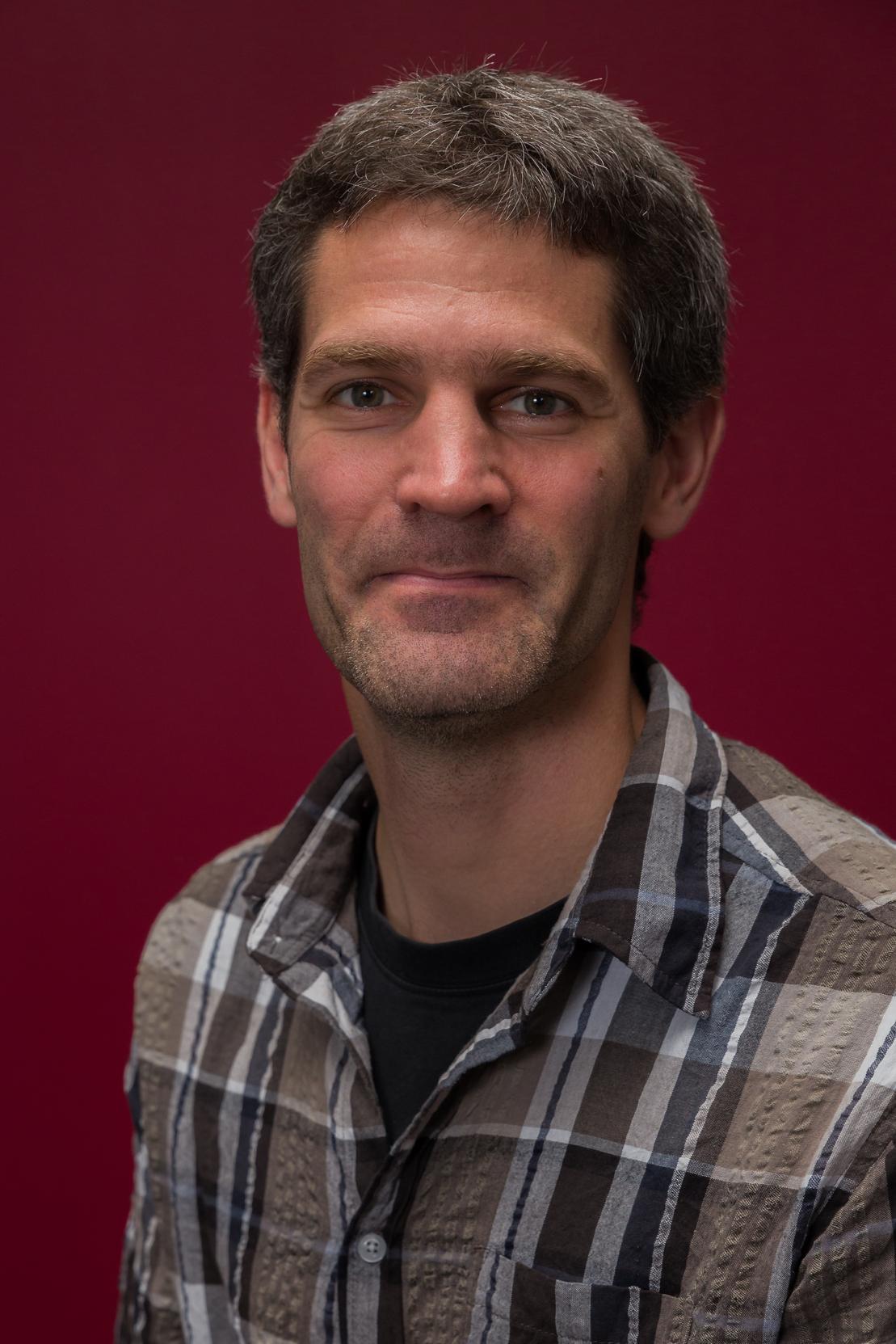 Porträt eines Mannes mit kurzem, grauem Haar, der ein kariertes Hemd trägt, vor einem roten Hintergrund.