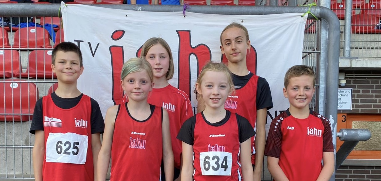 Sechs Kinder in roten Sporttrikots stehen vor einem Banner, jedem Kind ist eine Startnummer zugeordnet.
