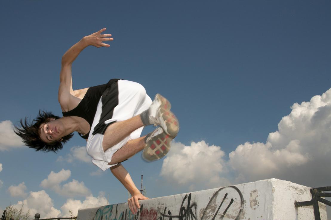 Junge führt einen akrobatischen Sprung über eine betonte Mauer unter blauem Himmel mit Wolken aus.