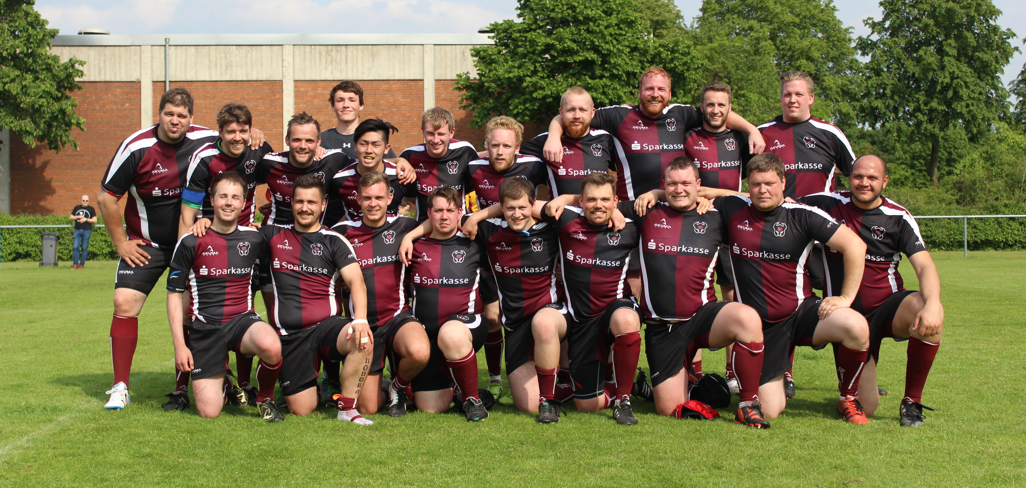 Gruppiertes Rugby-Team in schwarz-roten Trikots posiert l&auml;chelnd auf einem gr&uuml;nen Rasenfeld unter blauem Himmel.