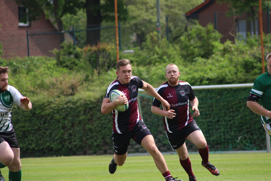Ein Rugbyspieler l&auml;uft mit dem Ball &uuml;ber ein gr&uuml;nes Feld, verfolgt von zwei anderen Spielern.