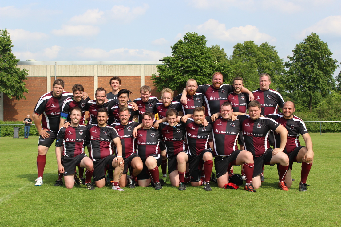 Gruppiertes Rugby-Team in schwarz-roten Trikots posiert l&auml;chelnd auf einem gr&uuml;nen Rasenfeld unter blauem Himmel.