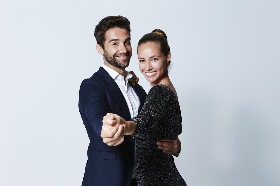 Paar in eleganter Kleidung l&auml;chelt und tanzt vor einem hellblauen Hintergrund, H&auml;nde ineinander.