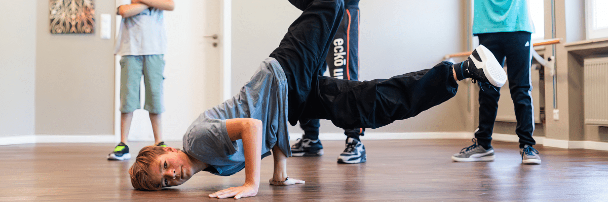 Junge in grauem T-Shirt führt einen Breakdance-Move auf dem Boden aus, umgeben von Zuschauern in sportlicher Kleidung.