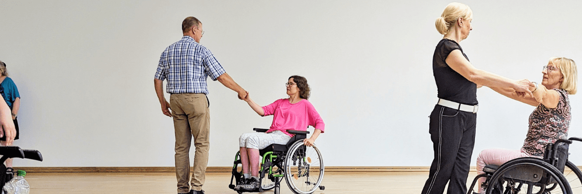 Tanzende Menschen, darunter zwei Rollstuhlfahrerinnen, in einem hellen Raum mit Holzfußboden.