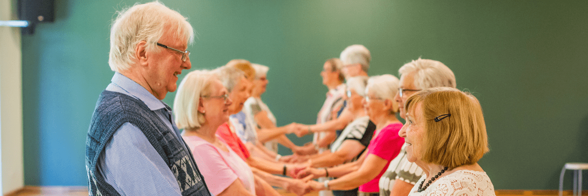 &Auml;ltere Menschen stehen in zwei Reihen und halten sich an den H&auml;nden in einem hellen Veranstaltungsraum.