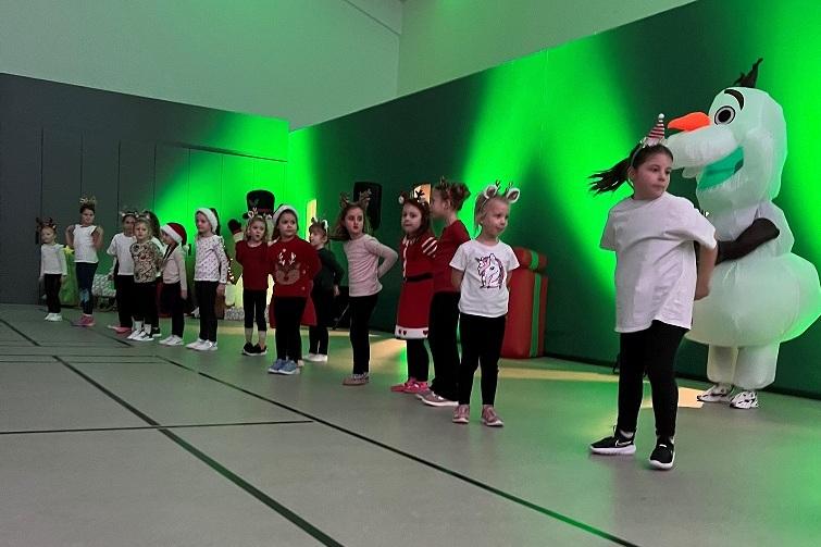 Gruppe von Kindern in festlicher Kleidung steht auf einer Sporthalle mit gr&uuml;nem Licht, ein Olaf-Kost&uuml;m ist sichtbar.