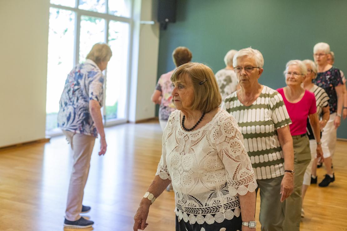Gruppe &auml;lterer Frauen tanzt in einem hellen Raum mit Holzboden und gro&szlig;en Fenstern.