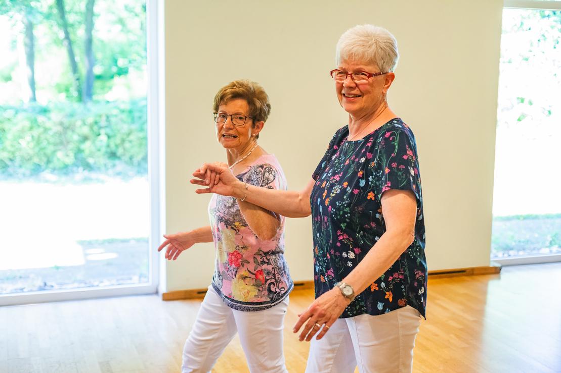 Zwei &auml;ltere Frauen tanzen l&auml;chelnd in einem hellen Raum mit gro&szlig;en Fenstern und Blick ins Gr&uuml;ne.