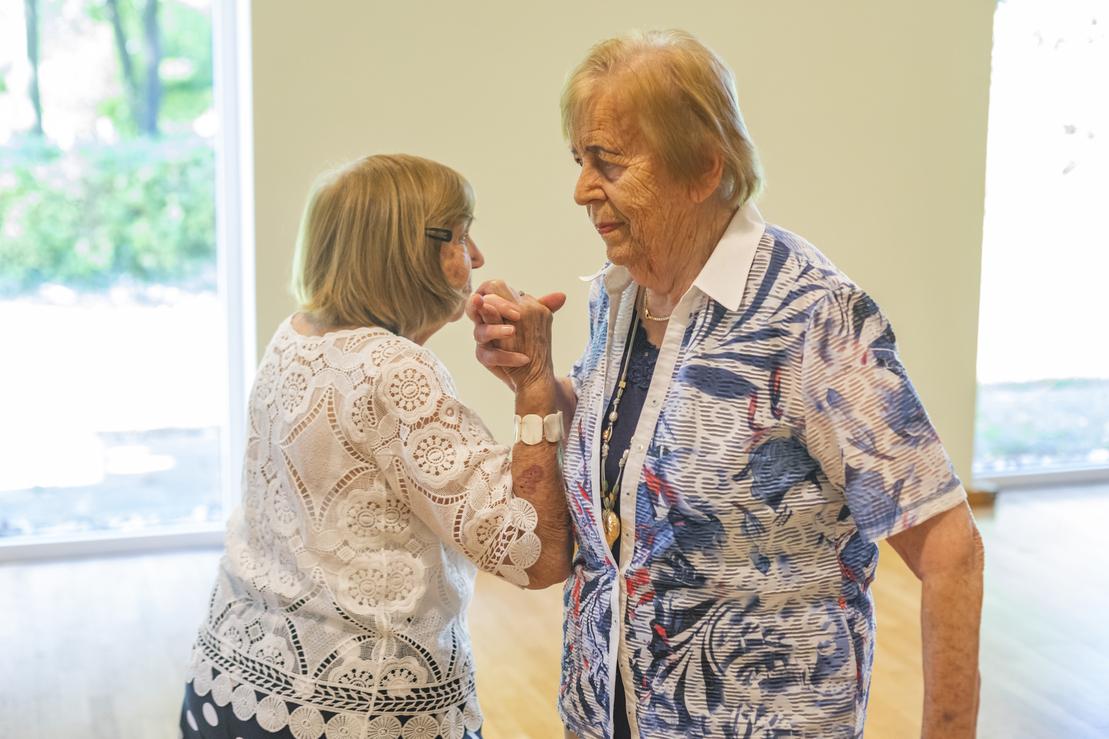 Zwei &auml;ltere Frauen tanzen miteinander in einem hellen Raum mit gro&szlig;en Fenstern.