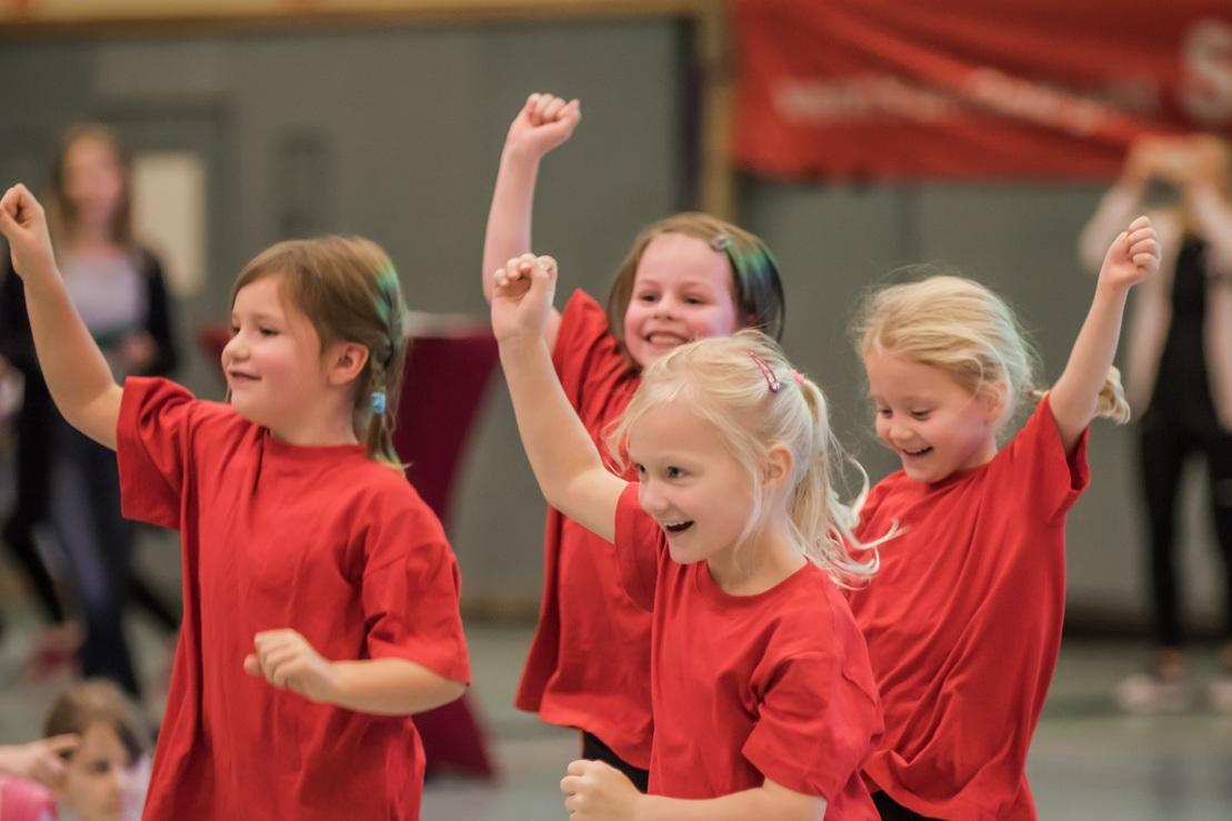 Vier Mädchen in roten T-Shirts tanzen und lächeln fröhlich in einer Sporthalle.