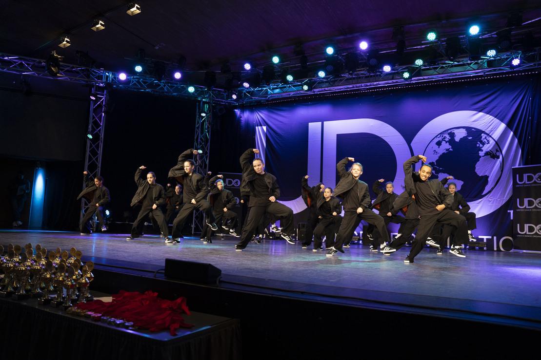 Tanzgruppe in schwarzen Outfits f&uuml;hrt energetischen Auftritt auf einer B&uuml;hne mit bunten Lichtern und Pokalen im Vordergrund vor.