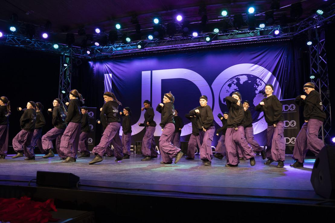 Gruppe von T&auml;nzern in Schwarz und Lila auf B&uuml;hne, vor einem Banner mit dem Logo der International Dance Organization (IDO).