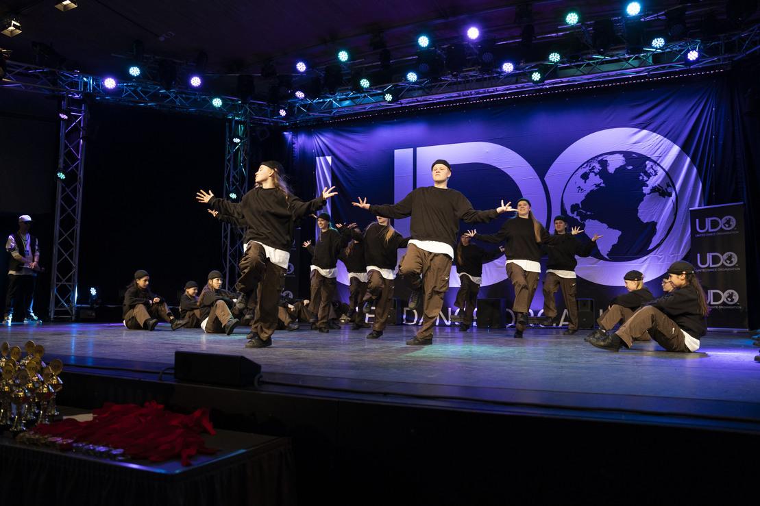 Tanzgruppe in schwarzen Outfits f&uuml;hrt eine Choreografie auf einer B&uuml;hne mit UDO-Logos und farbigen Lichtern aus.