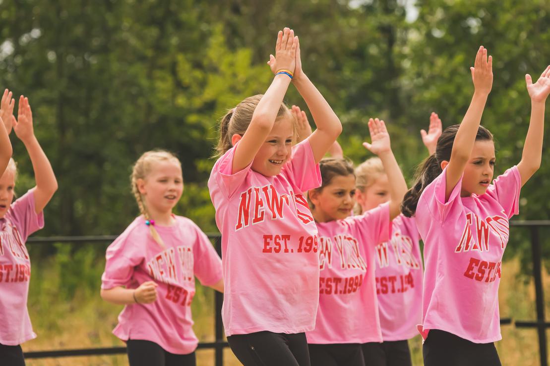 Gruppe von Kindern in pinken T-Shirts, die synchron in einer Aufführung mit erhobenen Händen tanzen.