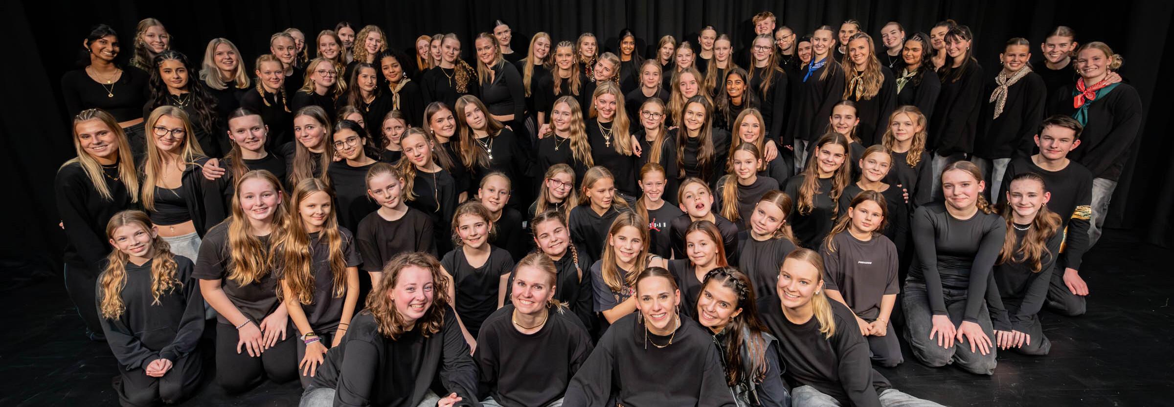 Gruppenportrait von &uuml;ber 70 T&auml;nzern in Schwarz auf einer B&uuml;hne mit schwarzem Hintergrund.