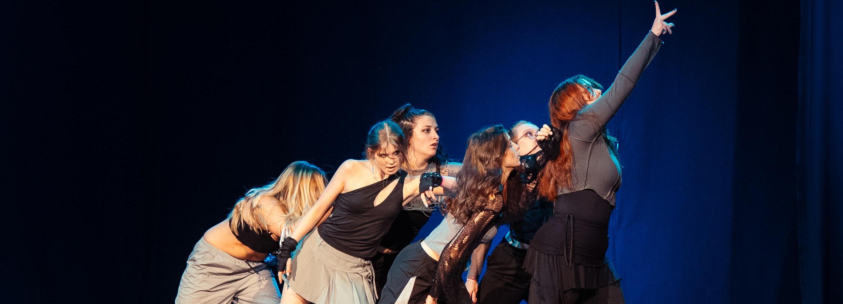 Sechs T&auml;nzerinnen in schwarzer und grauer Kleidung geben eine synchronisierte Performance auf einer B&uuml;hne.