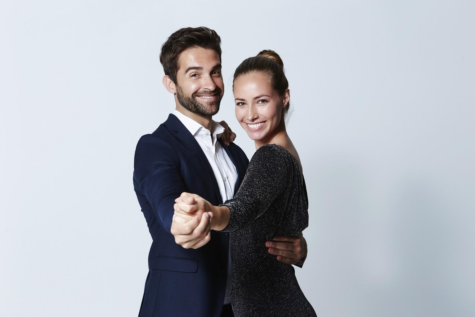 Ein Mann und eine Frau tanzen lächelnd zusammen in eleganter Kleidung vor einem hellen Hintergrund.