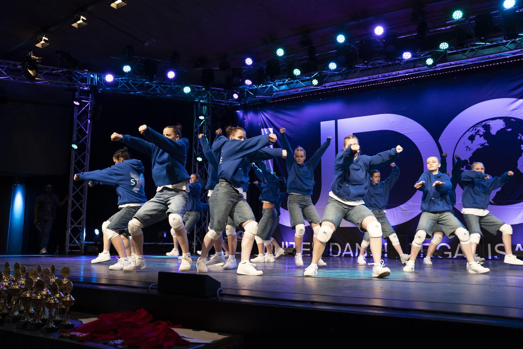 Tanzgruppe in blauen Hoodies und Shorts f&uuml;hrt eine choreografierte Auff&uuml;hrung auf einer B&uuml;hne mit Lichtern aus.